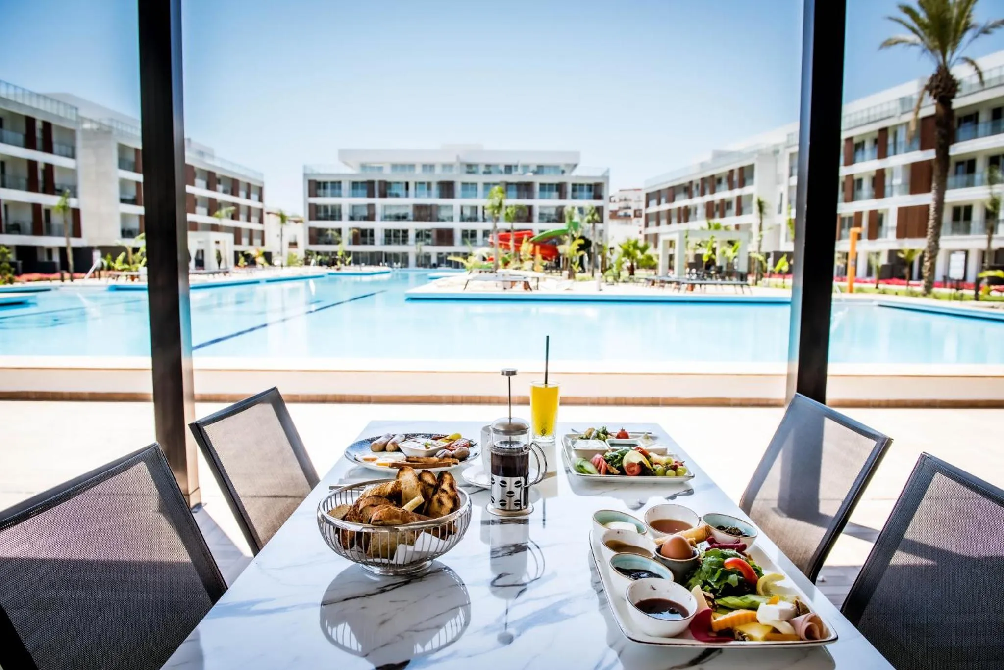 Une table est dressée avec un petit-déjeuner face à une piscine et à des bâtiments.