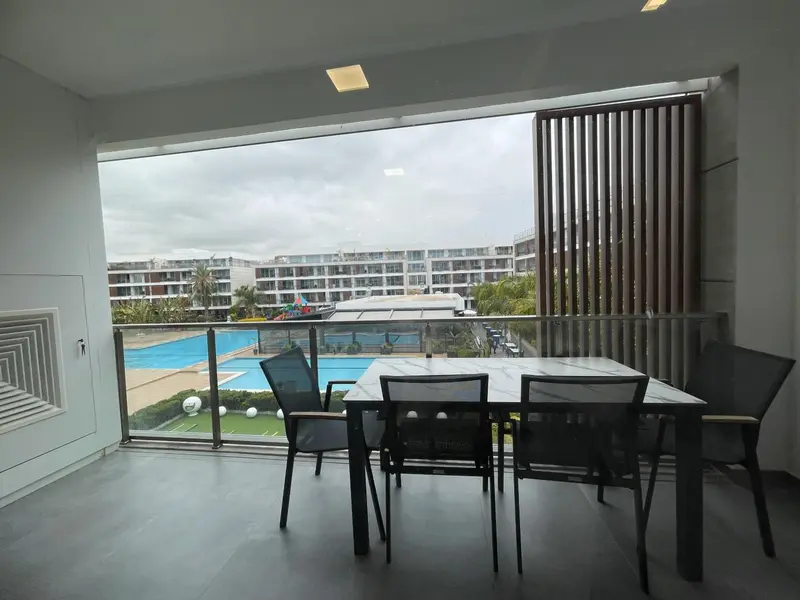 Terrasse avec table et chaises donnant sur une piscine et des bâtiments.