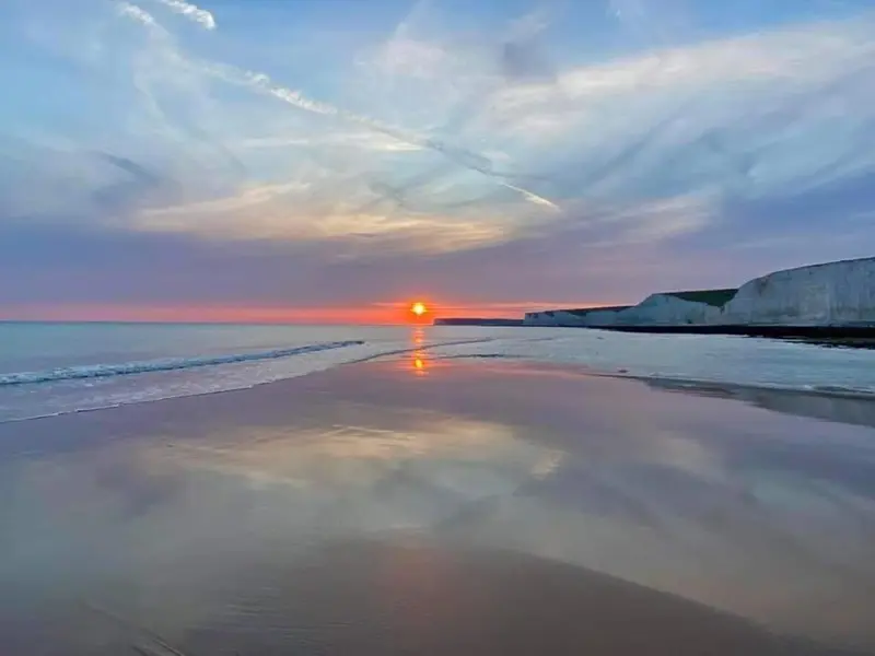 Coucher de soleil sur une plage de sable avec une mer calme et des falaises en arrière-plan.