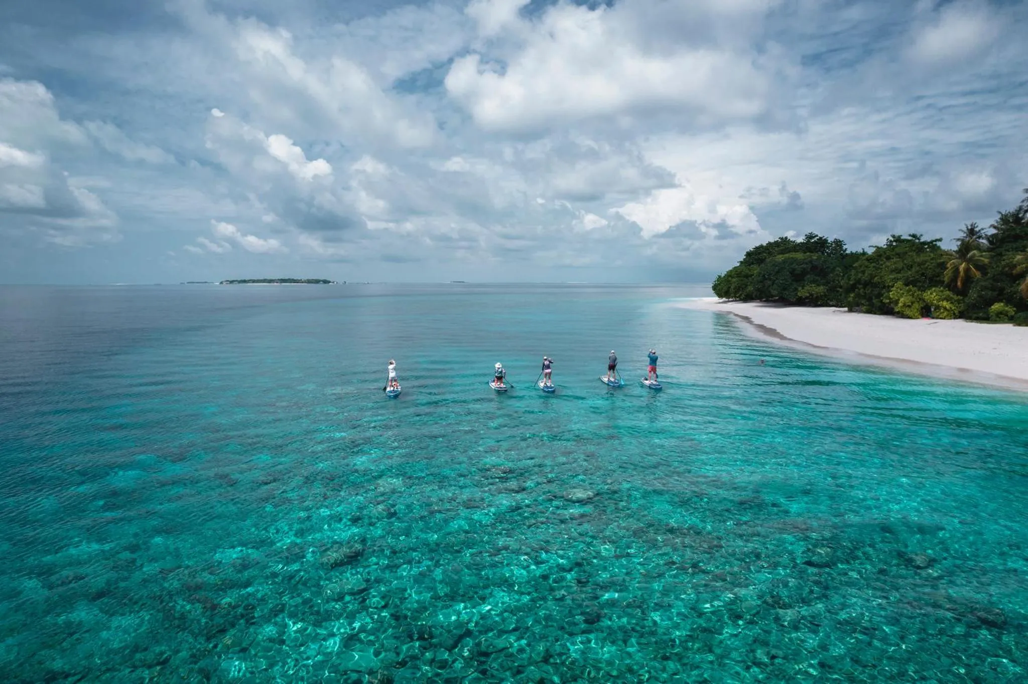 Vue aérienne d'une plage de sable blanc et de personnes pratiquant le stand-up paddle dans une eau turquoise.