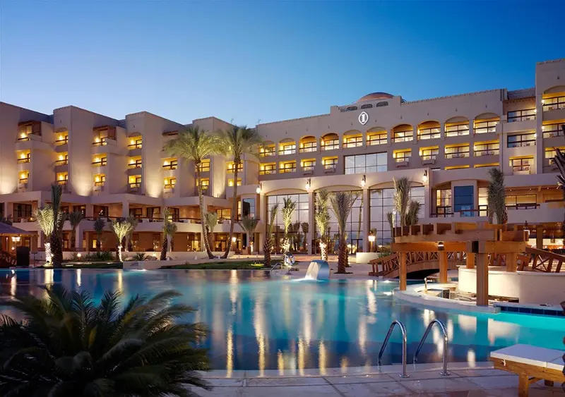 Un bâtiment beige illuminé surplombe une piscine turquoise au crépuscule.