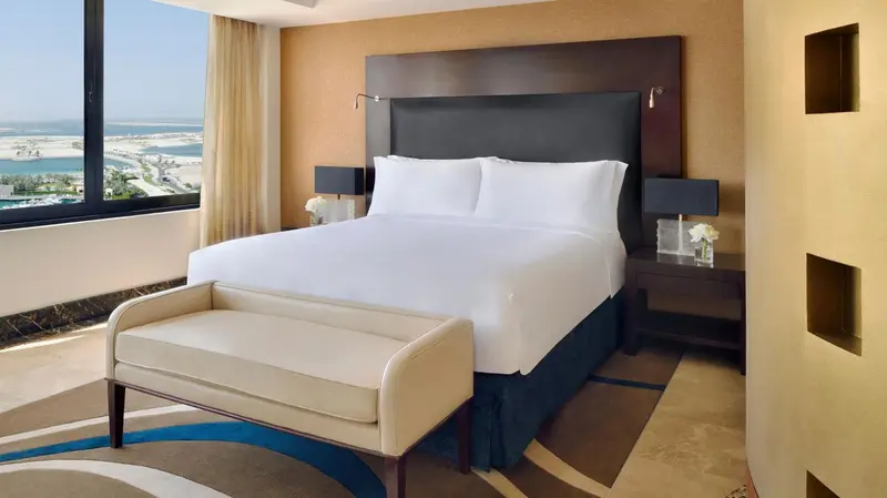 Une chambre d'hôtel lumineuse avec un grand lit et un banc au pied du lit.