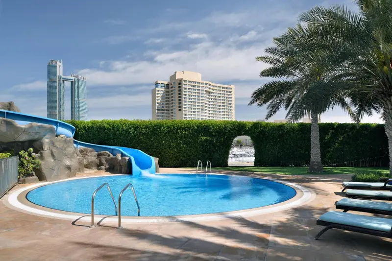 Une piscine avec toboggan et transats, devant un bâtiment et une végétation luxuriante.