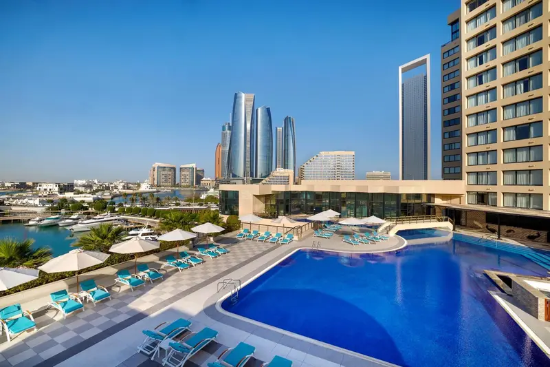 Une piscine turquoise bordée de transats bleus offre une vue sur un paysage urbain moderne.