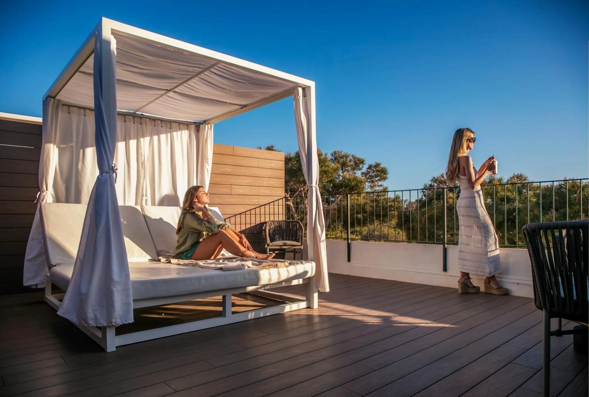Deux femmes se détendent sur une terrasse ensoleillée avec un canapé et une vue sur la végétation.