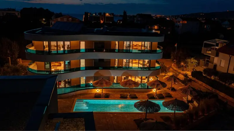Bâtiment moderne éclairé avec piscine et parasols.