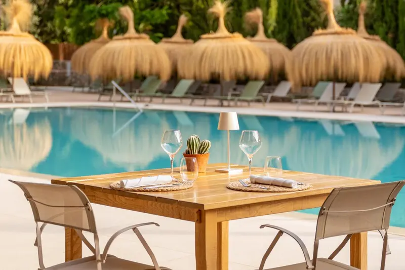 Table dressée au bord d'une piscine avec des parasols en chaume en arrière-plan.