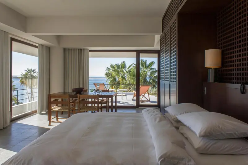 Une chambre lumineuse avec un lit blanc et une table à manger près d'une baie vitrée donnant sur la mer.