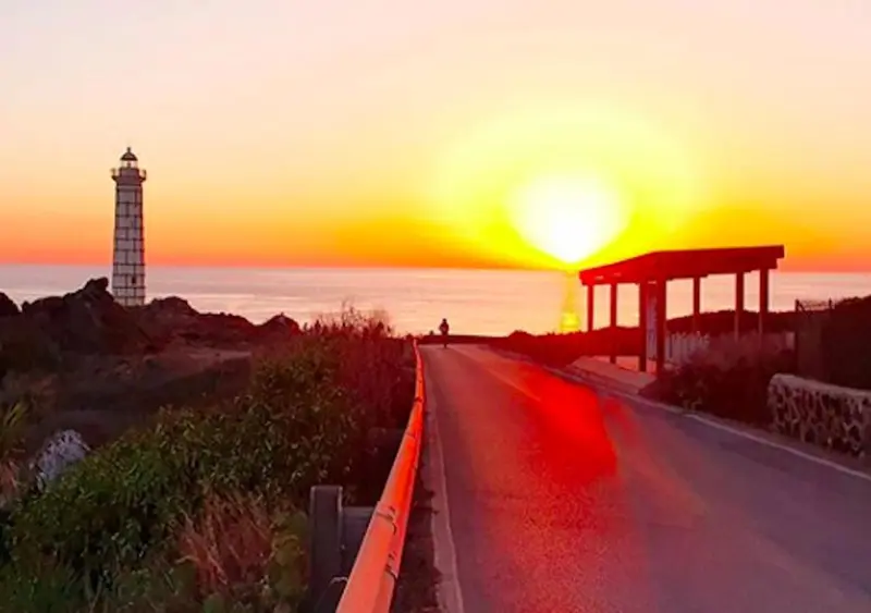 Une route côtière mène vers un phare au coucher du soleil.