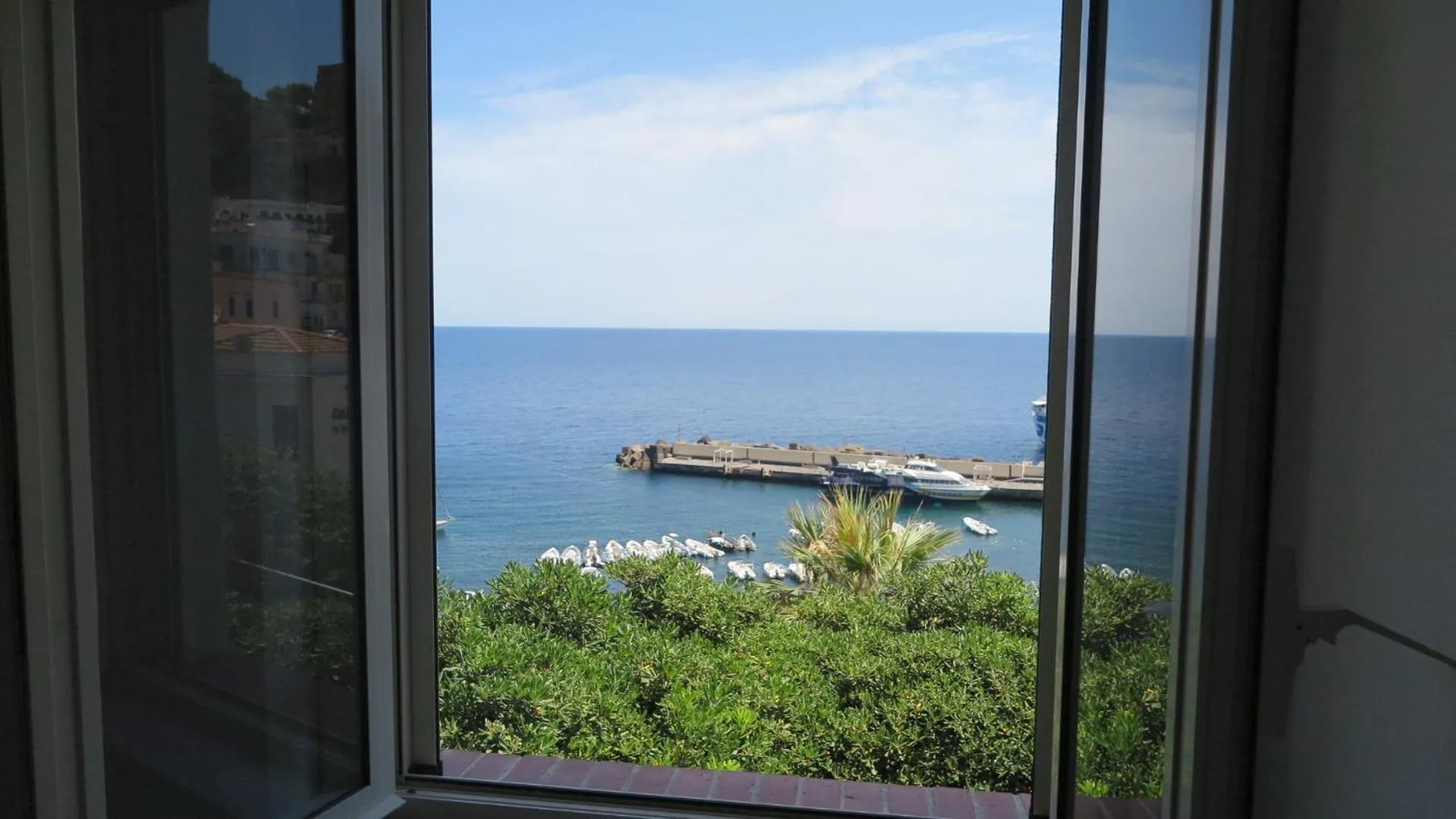 Vue sur la mer depuis une fenêtre ouverte, avec un port et une végétation luxuriante.