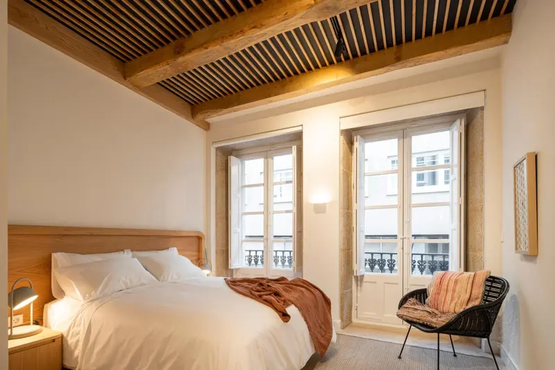 Une chambre lumineuse avec un lit blanc et un fauteuil noir près de fenêtres.