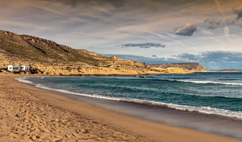 Une plage de sable fin borde une côte rocheuse sous un ciel nuageux.