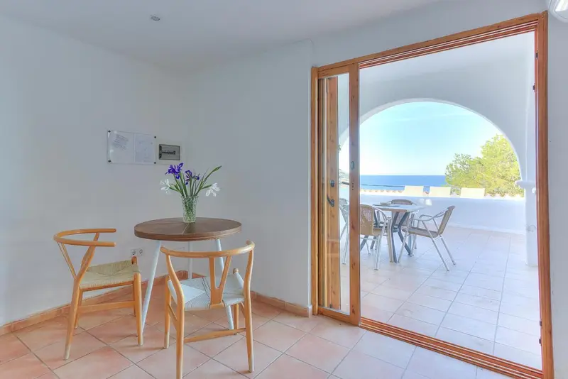 Une petite table ronde et deux chaises en bois se trouvent devant une baie vitrée ouverte sur une terrasse.