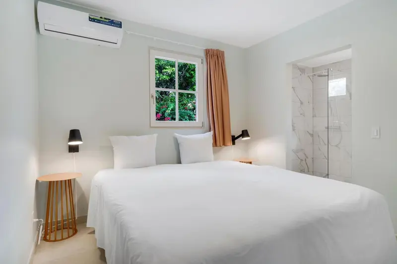 Une chambre lumineuse avec un lit blanc et une salle de bain carrelée.