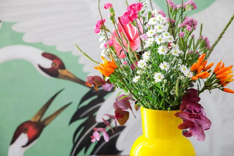 Un bouquet de fleurs multicolores dans un vase jaune se trouve devant un mur décoré d'oiseaux stylisés.