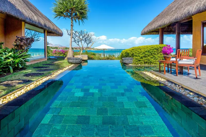 Une piscine rectangulaire turquoise s'étend devant des bâtiments aux toits de chaume.