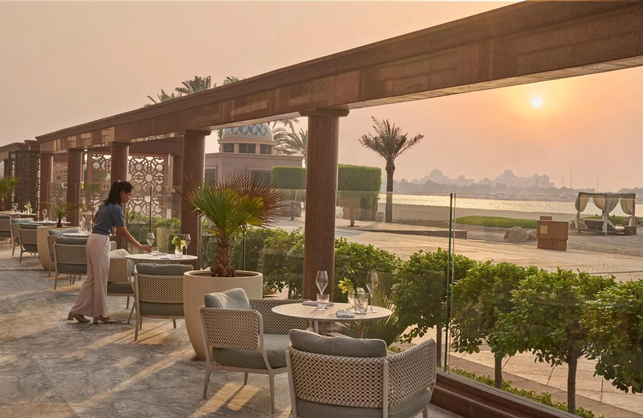 Une terrasse ombragée avec des tables et des chaises face à la mer au coucher du soleil.