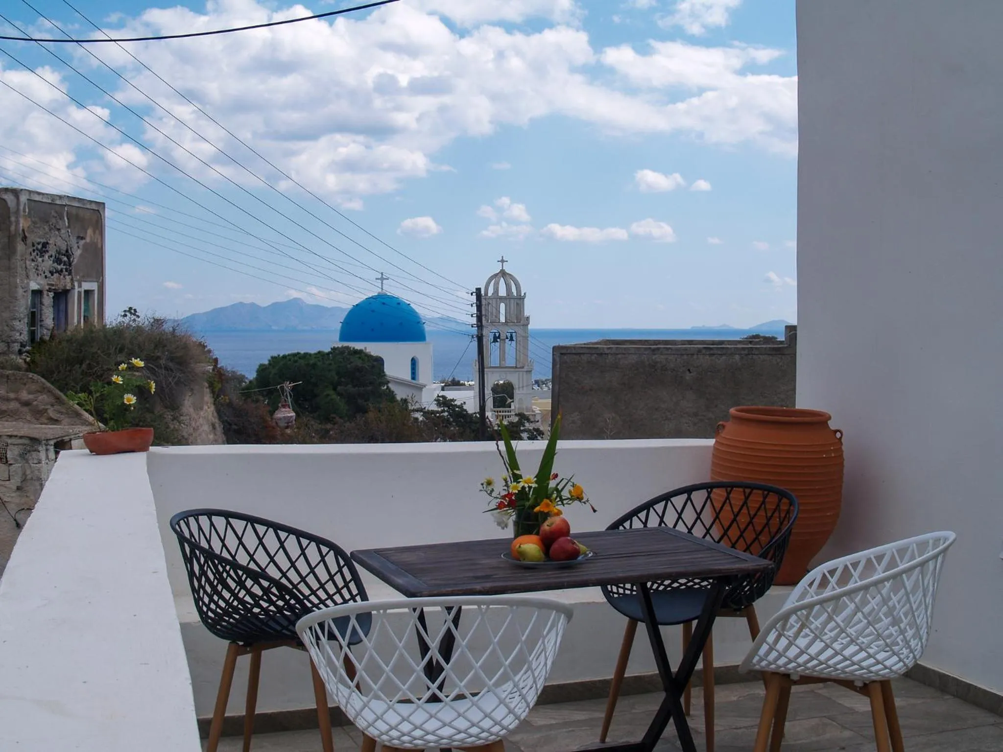 Une table et des chaises en osier sont disposées sur une terrasse avec vue sur la mer et un village.