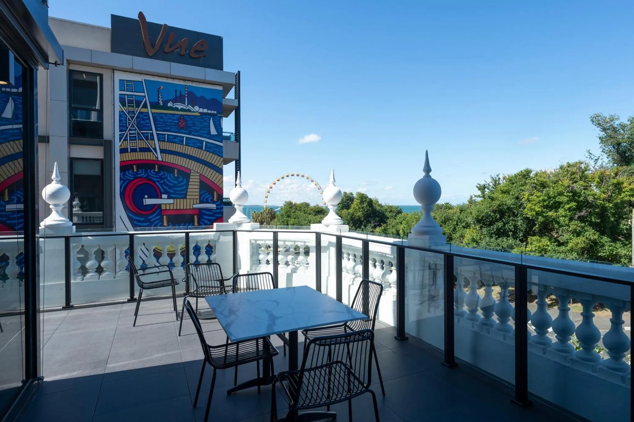 Terrasse aménagée avec tables et chaises offrant une vue sur la mer et une grande roue.