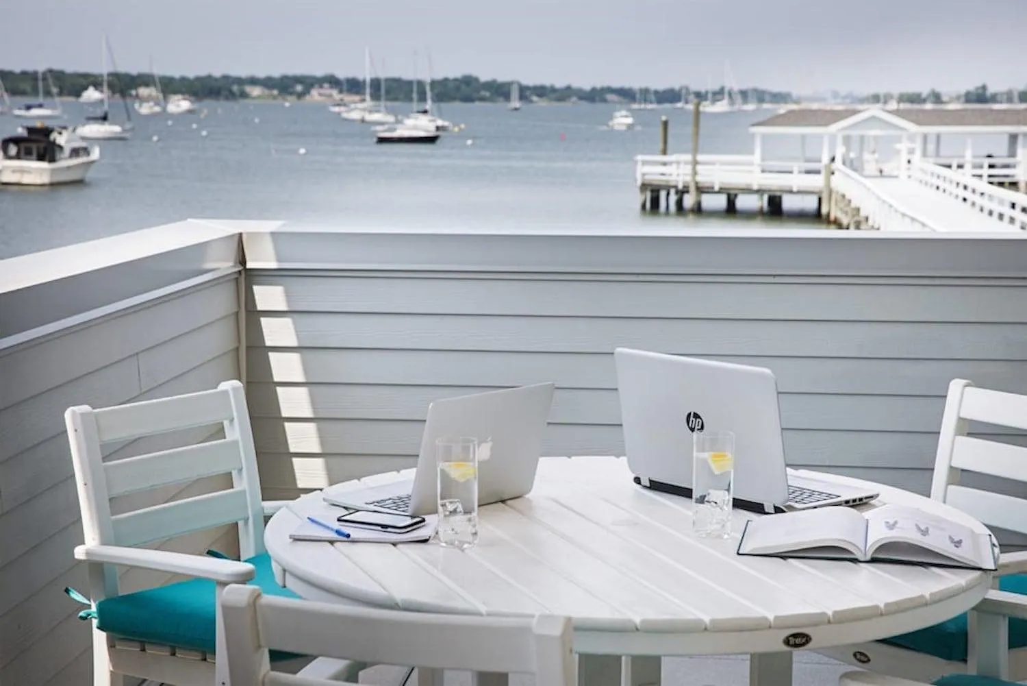 Table et chaises blanches sur une terrasse avec vue sur la mer et des bateaux.