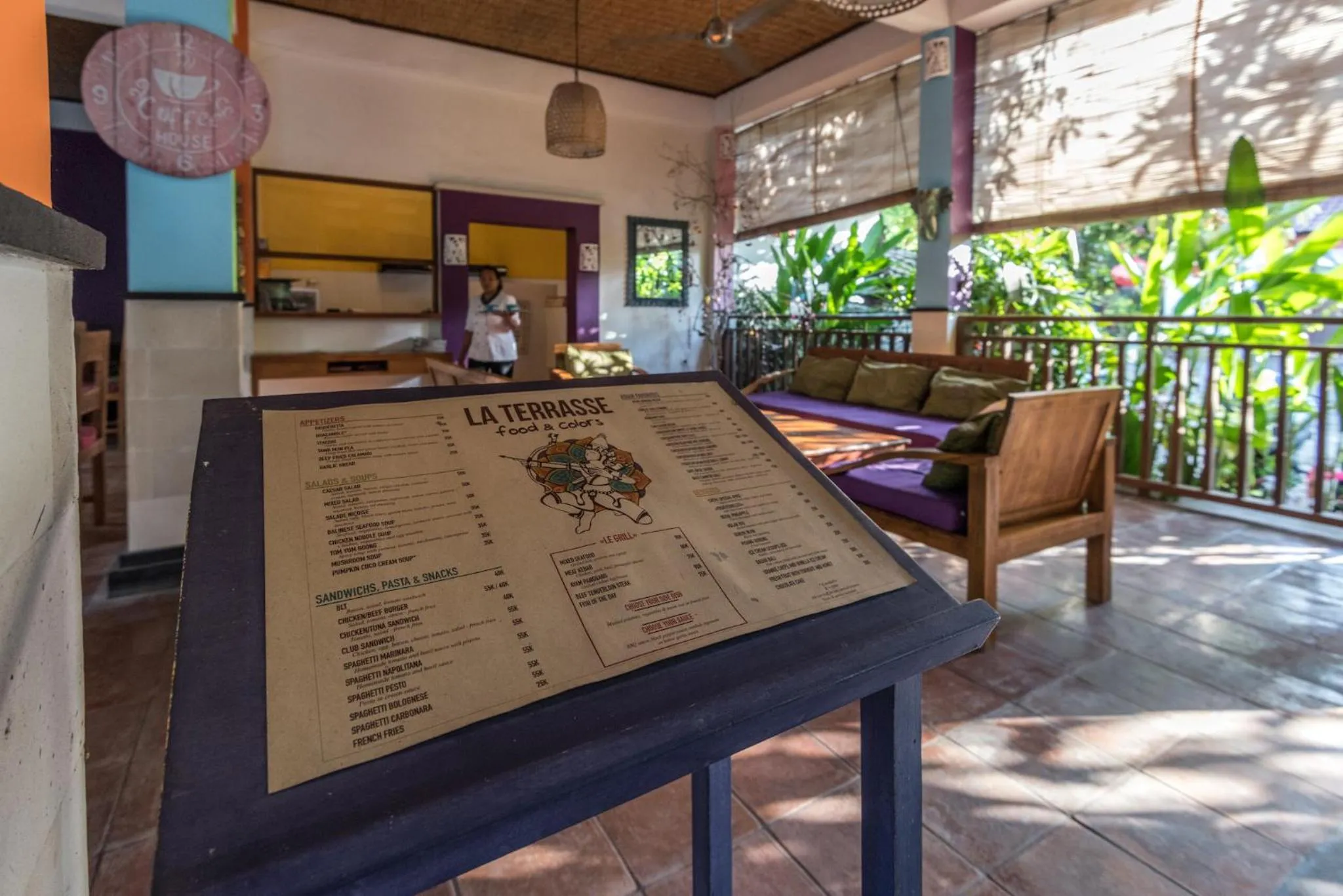 Un menu est présenté sur un chevalet devant une terrasse ombragée avec des tables et des canapés.
