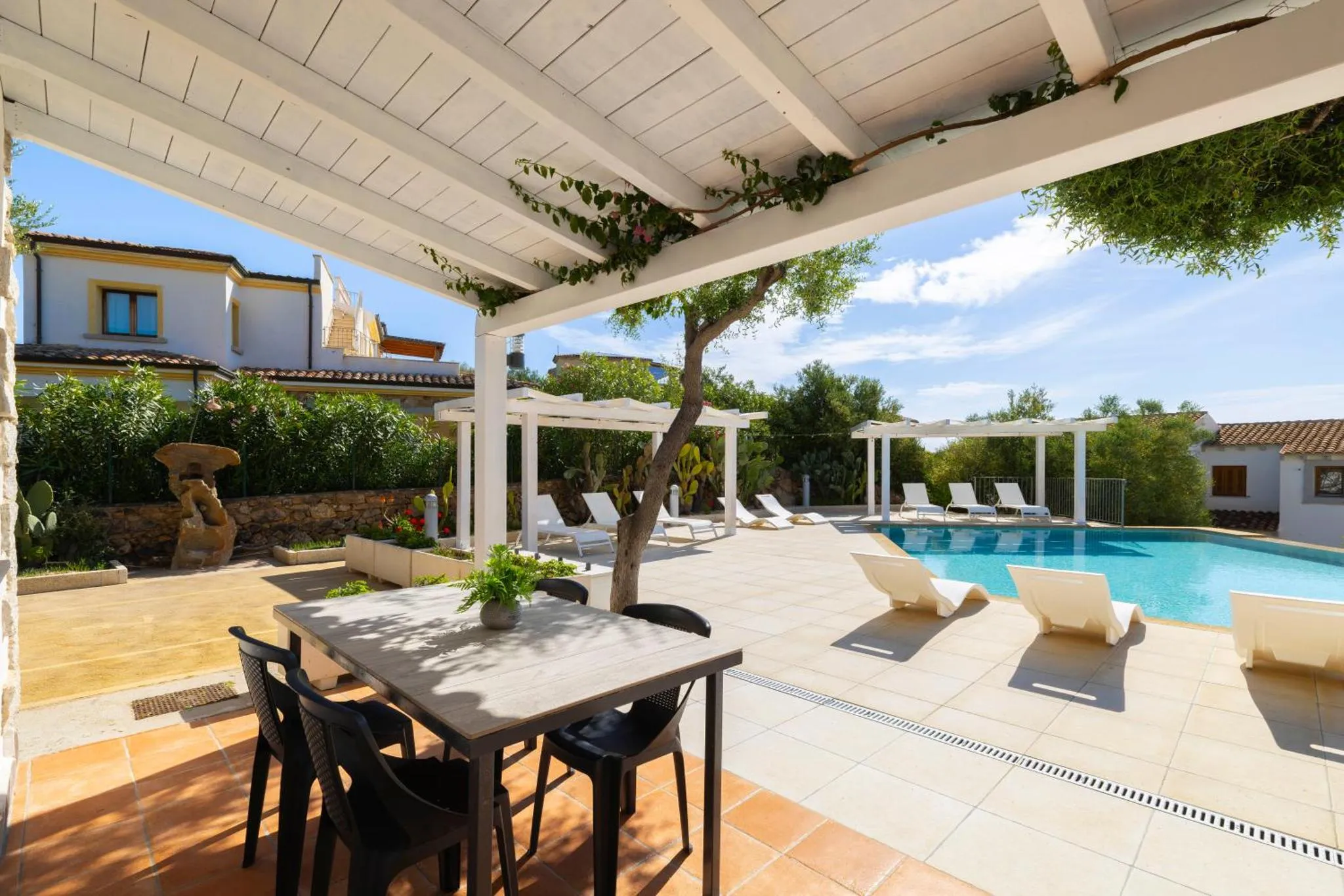 Terrasse ombragée avec table et chaises donnant sur une piscine et un espace de détente.