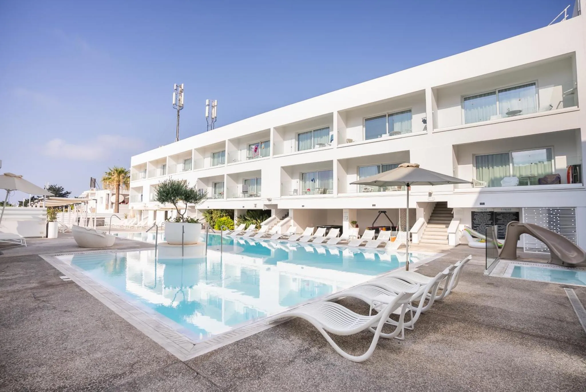 Piscine rectangulaire avec transats blancs et toboggan, devant un bâtiment blanc à balcons.