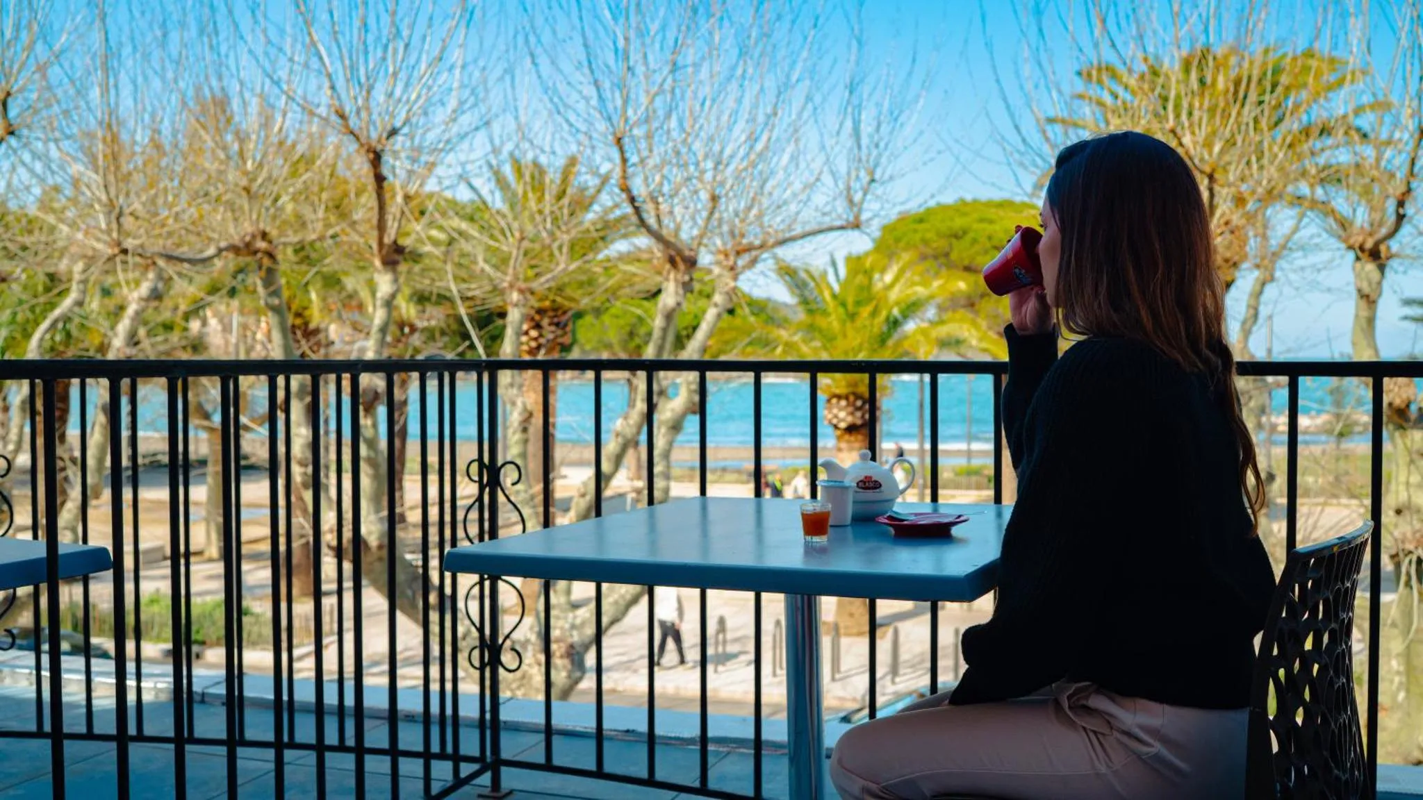 Une femme assise à une table en terrasse, face à la mer.