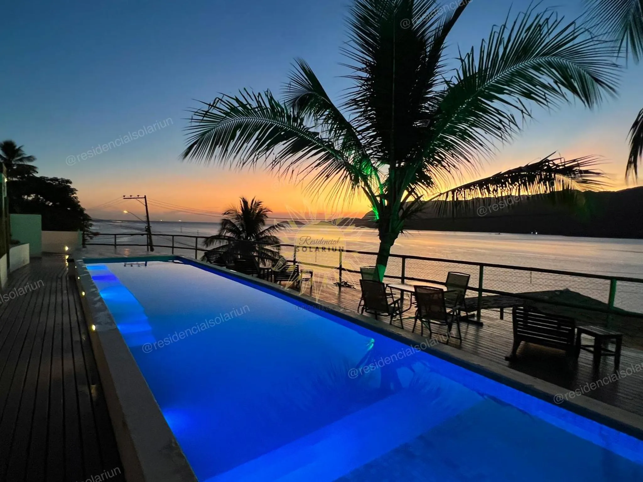 Piscine illuminée avec transats et palmiers au coucher du soleil.