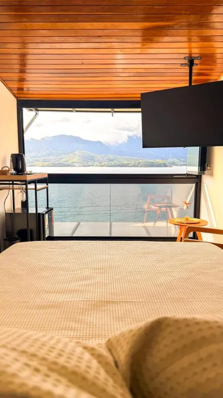 Une chambre lumineuse avec un lit face à une grande baie vitrée donnant sur un paysage montagneux et lacustre.