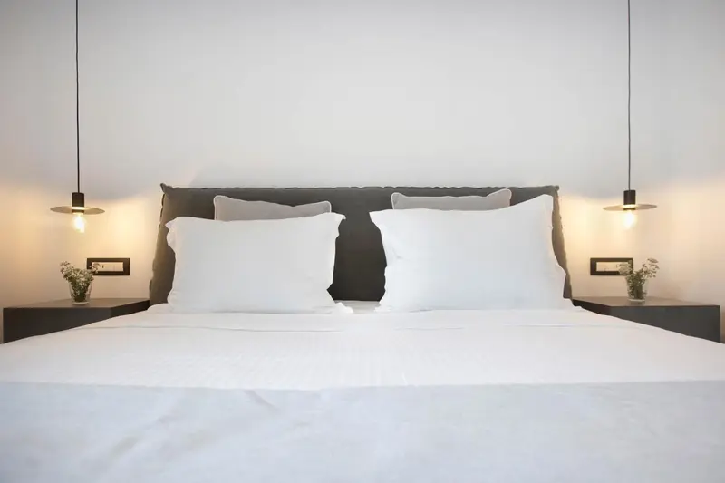 Un lit double avec des oreillers blancs et une tête de lit sombre est éclairé par deux lampes suspendues.