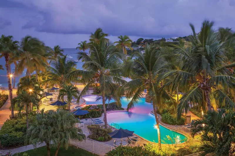Vue aérienne d'une piscine illuminée au crépuscule, entourée de végétation tropicale et donnant sur la mer.