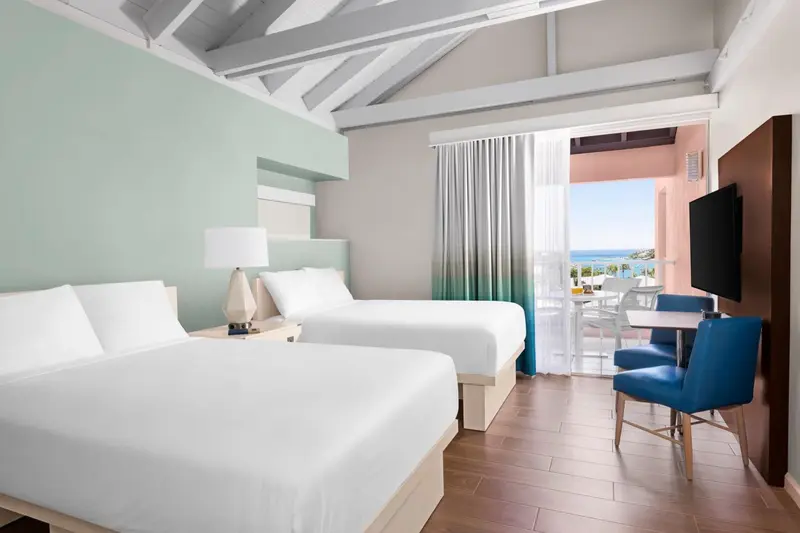 Chambre lumineuse avec deux lits et vue sur la mer depuis une terrasse.