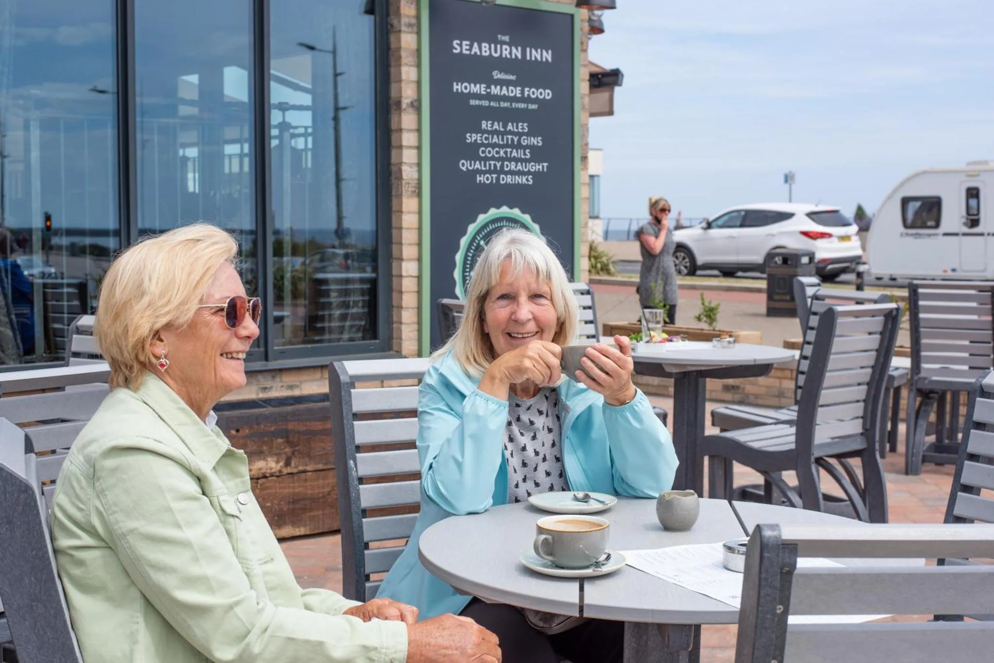 Deux femmes souriantes sont assises à une table en terrasse et dégustent une boisson.
