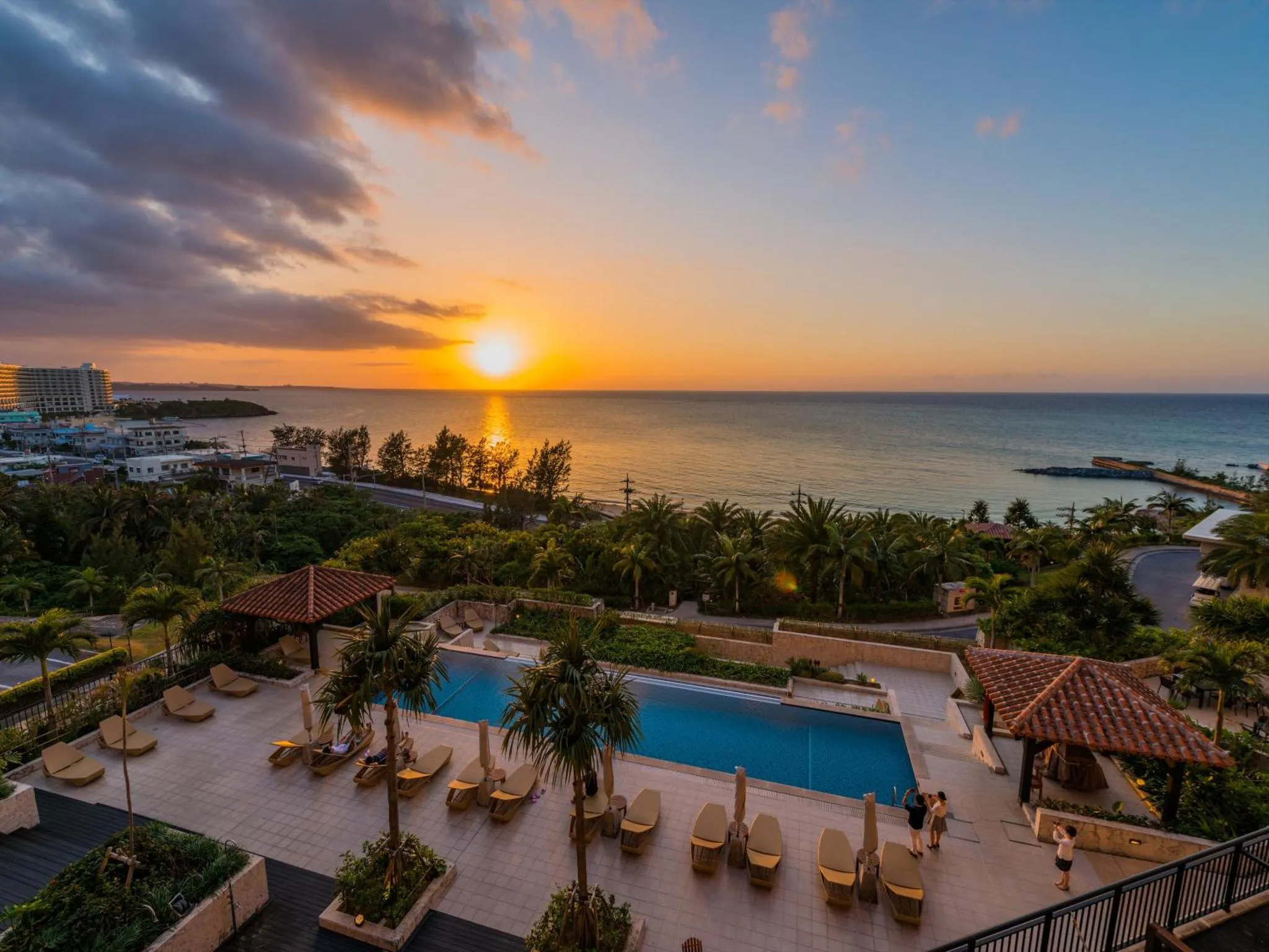 Une piscine avec des transats et des parasols, au premier plan, avec une vue sur la mer et un coucher de soleil.