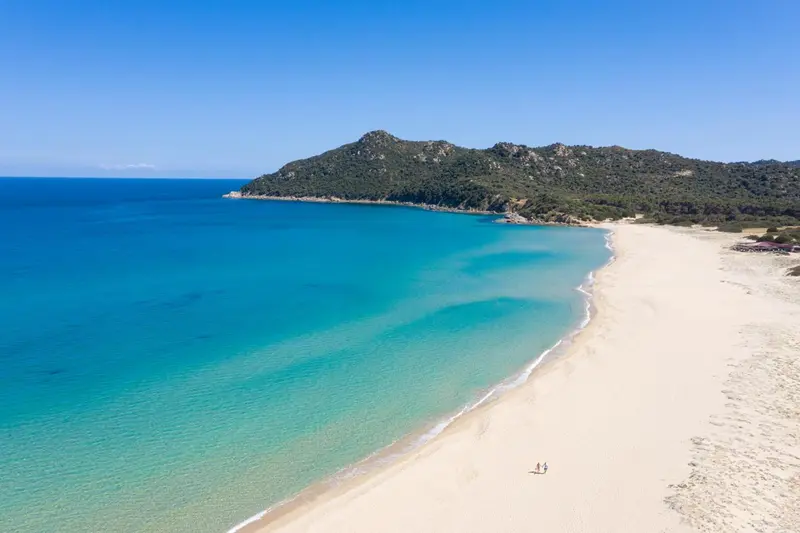 Vue aérienne d'une plage de sable fin bordée d'une eau turquoise et d'une colline verdoyante.