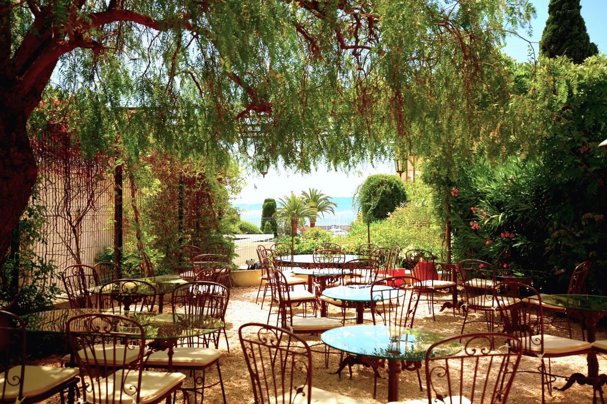 Terrasse ombragée avec tables et chaises en fer forgé, vue sur la mer en arrière-plan.