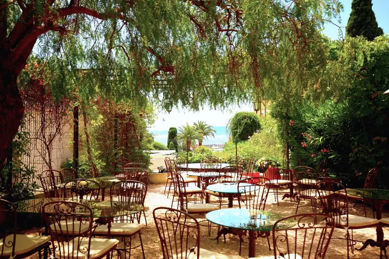 Terrasse ombragée avec tables et chaises en fer forgé, vue sur la mer en arrière-plan.
