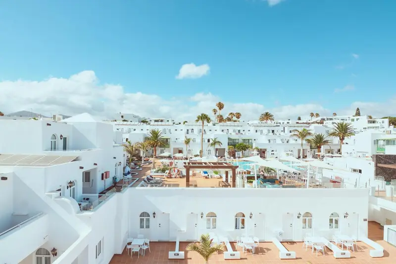 Vue aérienne d'un complexe hôtelier blanc avec piscine et palmiers sous un ciel bleu.
