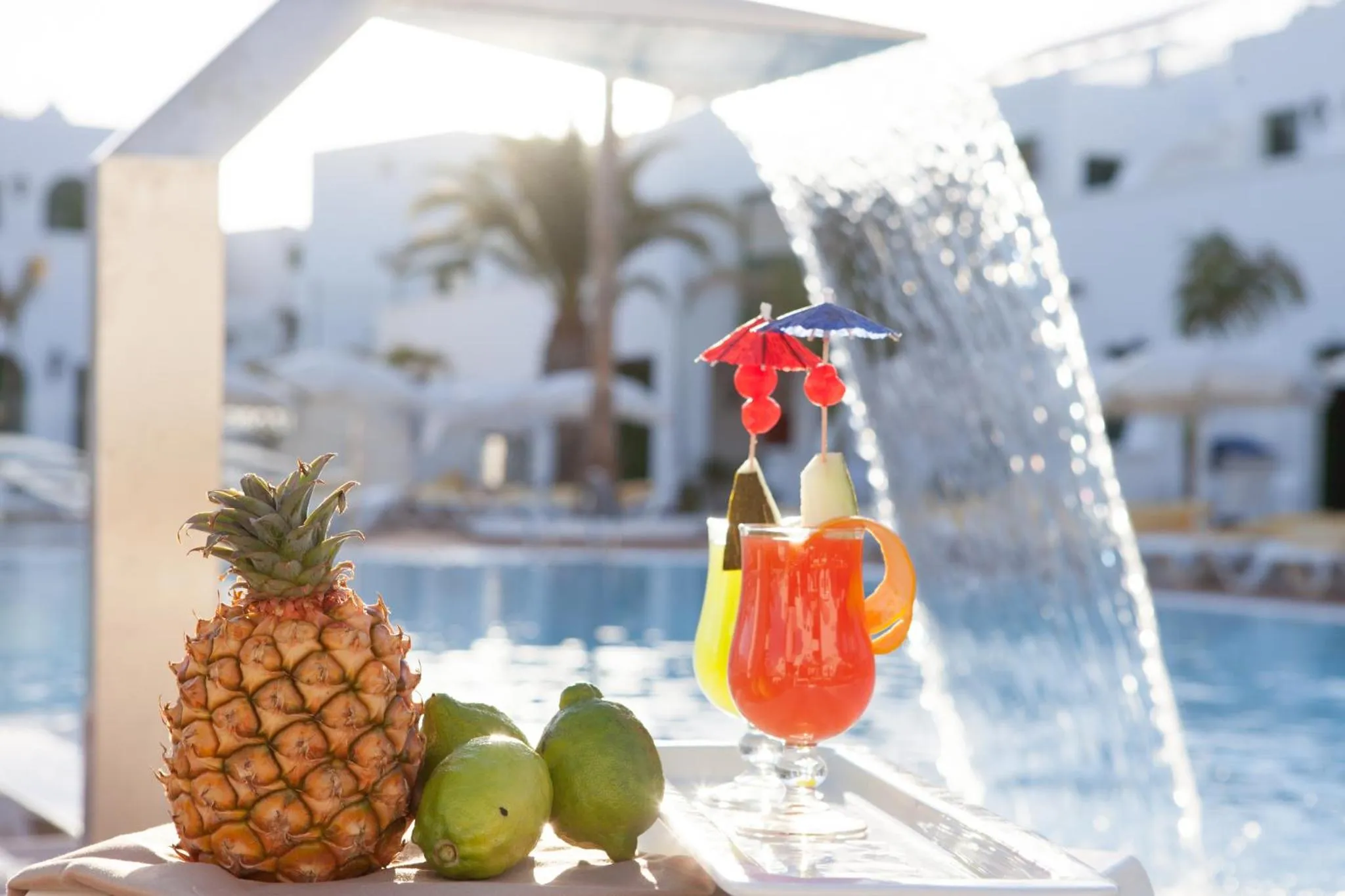 Un ananas, des citrons verts et un cocktail coloré sont présentés au bord d'une piscine.