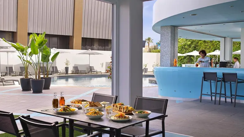 Une table est dressée devant un bar bleu turquoise, près d'une piscine ensoleillée.