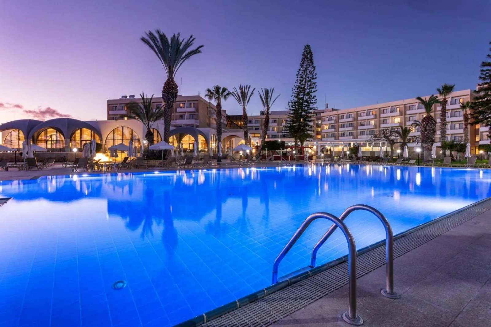 Piscine illuminée d'un hôtel au crépuscule, avec des palmiers en arrière-plan.