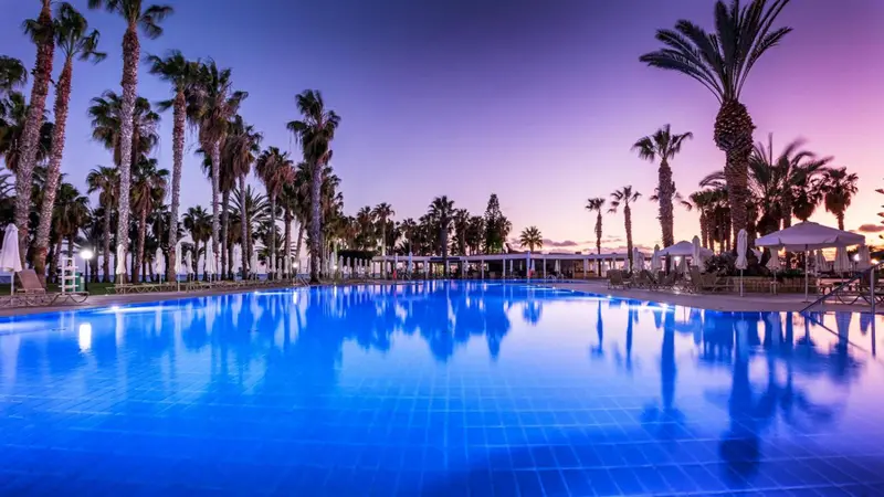 Une piscine illuminée reflète les palmiers et le ciel crépusculaire.