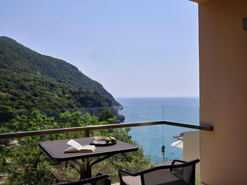 Une table avec un livre et des fruits est posée sur un balcon avec vue sur la mer et les montagnes.