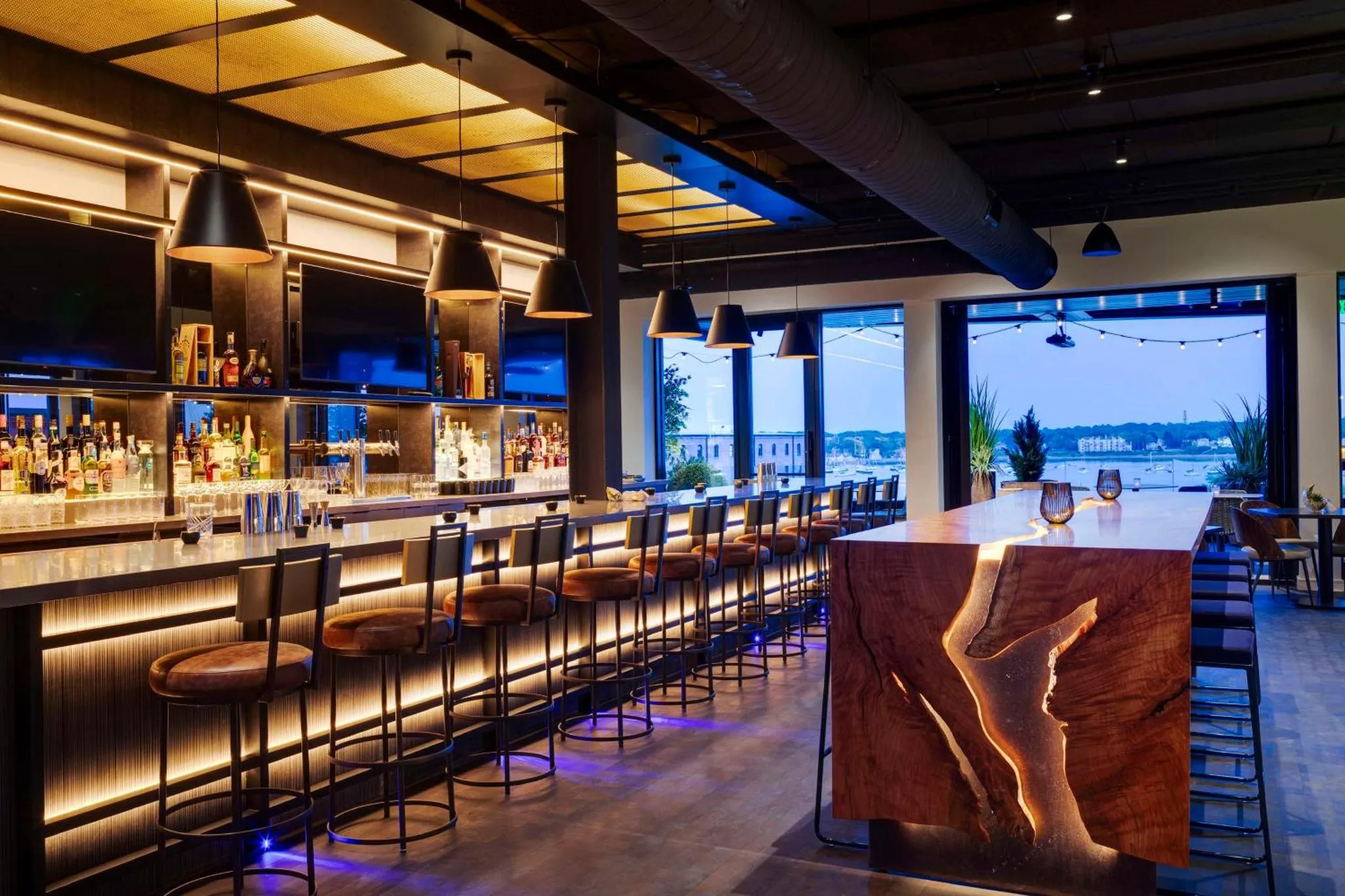 Un long comptoir de bar éclairé accueille des clients sur des tabourets hauts, avec une vue sur l'extérieur.