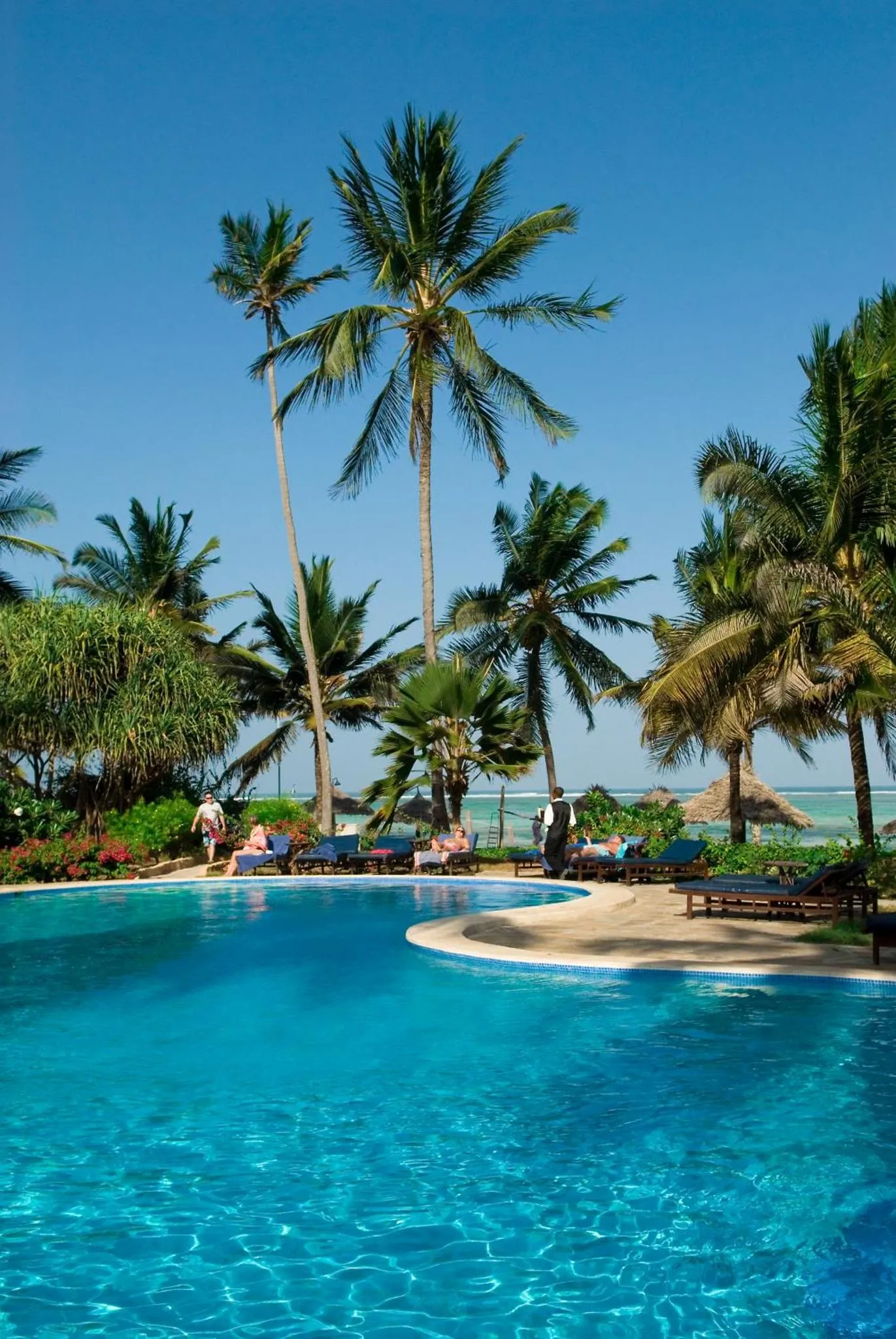 Une piscine turquoise bordée de transats, avec des palmiers et une plage en arrière-plan.
