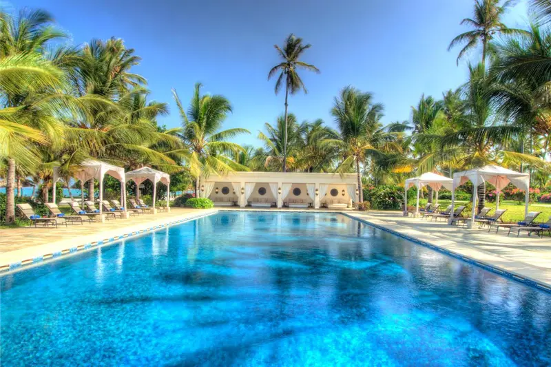 Une piscine bleue bordée de transats et de parasols, avec une végétation luxuriante en arrière-plan.