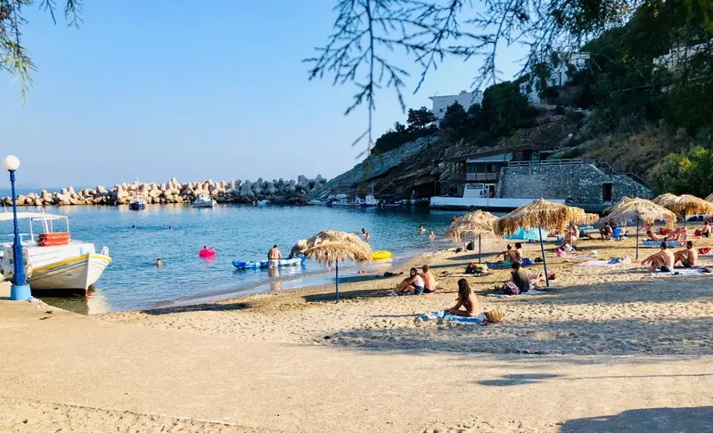 Une plage de sable avec des transats et des parasols, bordée par une mer bleue et un port.