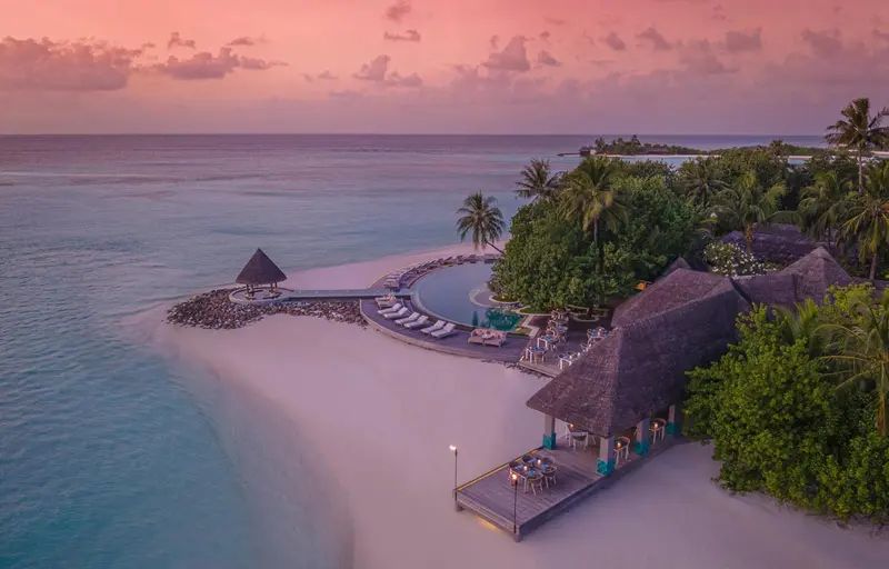 Vue aérienne d'une plage de sable blanc bordée de végétation tropicale et de bungalows sur pilotis.