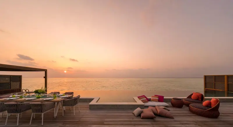 Terrasse en bois avec table à manger, piscine à débordement et mobilier d'extérieur face à la mer au coucher du soleil.